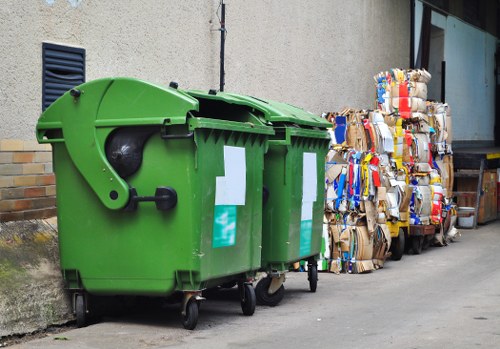 This image shows a bright green, large wheeled rubbish bin positioned on a concrete pavement, adjacent to a plain, pale grey building wall with small ventilation slots visible on the left side. The bin has a rounded, smooth surface with a closed lid and black wheels at the base, indicating it is designed for outdoor waste collection. To the right of the bin are multiple tightly packed bundles of flattened cardboard boxes, bound with twine, featuring various colours and printed logos, stacked along the sidewalk. In the background, part of a modern building with glass windows and a wooden frame can be seen. The scene appears to be in a commercial or industrial area, possibly in Croydon, where [officeclearancecroydon.co.uk](https://officeclearancecroydon.co.uk/) offers rubbish removal services. The overall setting is outdoors under natural daylight, with the waste items clearly visible and organized for collection or disposal.