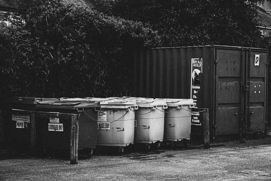 A large metal rubbish bin with a weathered, slightly corroded surface, situated outdoors on a paved pathway beside a grassy area with lush green foliage and trees in the background. The bin features a
